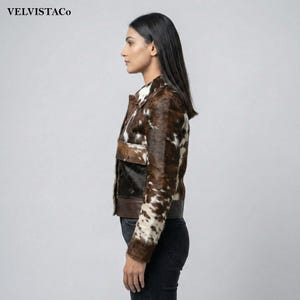 May include: A brown and white cowhide jacket with a collared neckline and two front pockets. The jacket has a dark brown leather trim and a black panel on the front. The brand name "VELVISTACO" is visible in the top left corner.