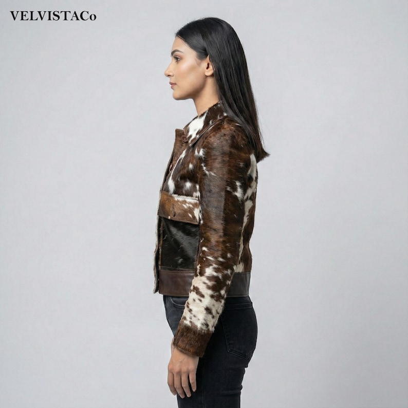 May include: A brown and white cowhide jacket with a collared neckline and two front pockets. The jacket has a dark brown leather trim and a black panel on the front. The brand name "VELVISTACO" is visible in the top left corner.