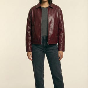 May include: A burgundy leather jacket with a collared neckline and a zipper closure. The jacket is paired with a gray t-shirt and dark gray jeans. The image also shows black ankle boots. The brand name "VELVISTACO" is visible in the top left corner.
