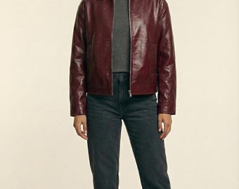 May include: A burgundy leather jacket with a collared neckline and a zipper closure. The jacket is paired with a gray t-shirt and dark gray jeans. The image also shows black ankle boots. The brand name "VELVISTACO" is visible in the top left corner.
