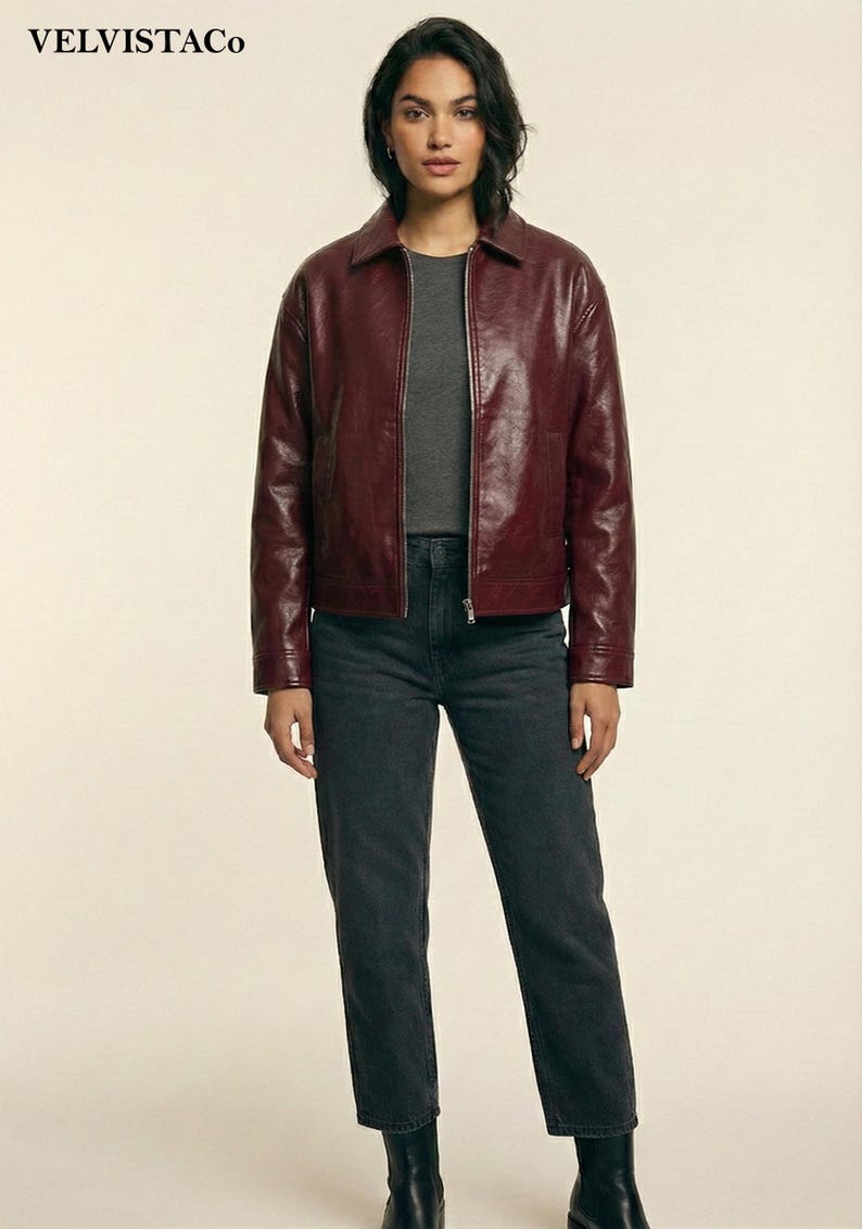May include: A burgundy leather jacket with a collared neckline and a zipper closure. The jacket is paired with a gray t-shirt and dark gray jeans. The image also shows black ankle boots. The brand name "VELVISTACO" is visible in the top left corner.