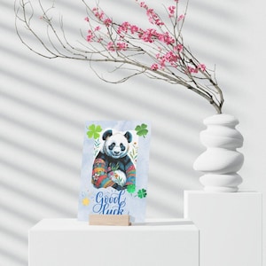 May include: A greeting card featuring a watercolor panda with a colorful sweater, surrounded by shamrocks and floral accents. The card has the words "Good Luck" in script. The card is displayed on a white stand.