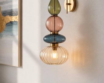 May include: A multi-colored glass wall sconce with a brass finish. The light fixture features stacked, rounded glass elements in green, pink, blue, and amber. The light bulb is visible inside the amber glass. The fixture is mounted on a white wall.