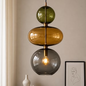 Op de afbeelding: Een hanglamp met drie gestapelde glazen bollen in olijfgroen, amber en antracietgrijs. De bollen zijn verbonden door een centrale messing stang, met een gloeiende lamp zichtbaar in de onderste antracietgrijze glazen bol.