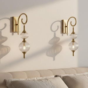 May include: Two gold-toned wall sconces with clear, ribbed glass globes. Each sconce has a decorative, curved arm and a rectangular base. The sconces are mounted on a light-colored wall, casting shadows.