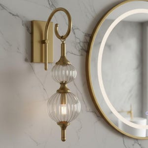 May include: A gold-toned wall sconce with two ribbed glass globes and a decorative loop. The sconce is mounted on a rectangular base against a marble-patterned wall. A round mirror with a gold frame is in the background.