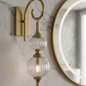 May include: A gold-toned wall sconce with a curved arm and two clear, ribbed glass globes. The fixture is mounted on a rectangular base and hangs near a round mirror with a gold frame. The light fixture is designed for indoor use.