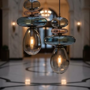 May include: Two pendant lights featuring stacked, translucent, smoke-colored glass elements in varying rounded shapes. Each fixture includes a clear, teardrop-shaped glass shade at the bottom housing a glowing filament bulb, suspended by thin black cords.