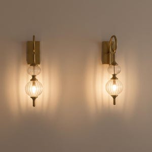 May include: Two wall sconces with a brass finish and clear, ribbed glass globes. Each sconce has a vertical rectangular backplate, a decorative loop, and a small, elongated base. The sconces are illuminated, casting a warm glow on the wall.