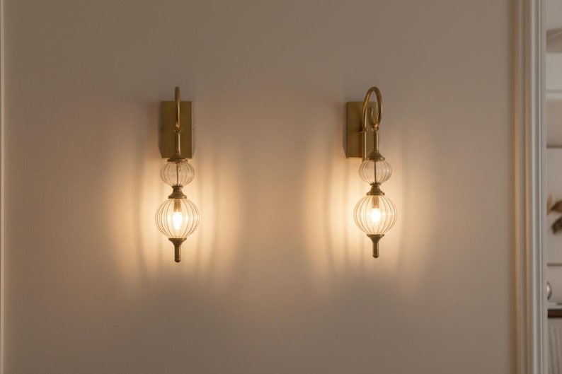 May include: Two wall sconces with a brass finish and clear, ribbed glass globes. Each sconce has a vertical rectangular backplate, a decorative loop, and a small, elongated base. The sconces are illuminated, casting a warm glow on the wall.
