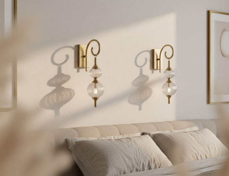 May include: Two gold-toned wall sconces with clear, ribbed glass globes. Each sconce has a decorative, curved arm and a rectangular base. The sconces are mounted on a light-colored wall, casting shadows.
