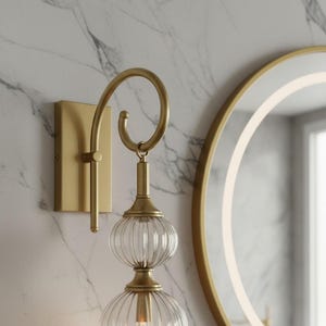 May include: A gold-toned wall sconce with a curved arm and two clear, ribbed glass globes. The sconce is mounted on a rectangular base against a marble-patterned wall, with a round mirror in the background.