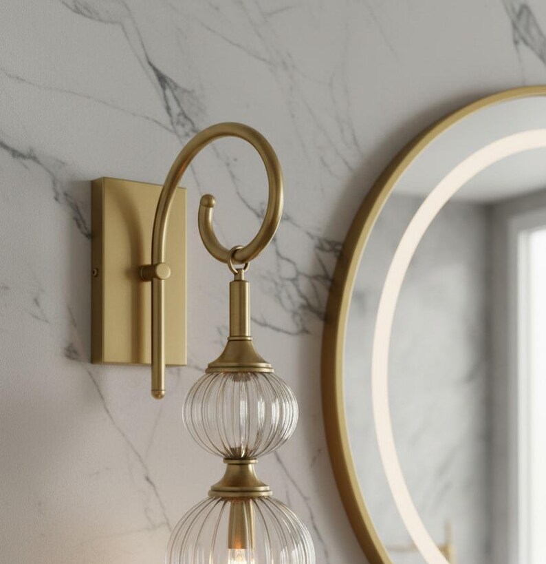 May include: A gold-toned wall sconce with a curved arm and two clear, ribbed glass globes. The sconce is mounted on a rectangular base against a marble-patterned wall, with a round mirror in the background.
