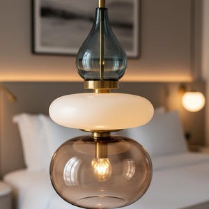 Pode incluir: Uma moderna luminária pendente com um design em camadas. Possui um topo de vidro azul em forma de gota, um disco branco e um globo de vidro castanho na parte inferior. A luminária tem detalhes em metal dourado e uma lâmpada visível.