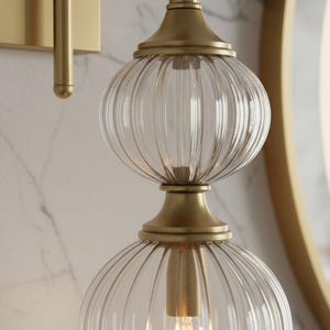 May include: A gold-toned wall sconce with a clear, ribbed glass shade. The sconce features a double-globe design, with a light bulb visible inside. The fixture is mounted on a marble-patterned wall, with a gold-framed mirror in the background.