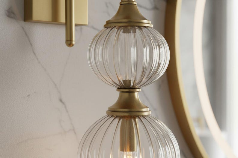 May include: A gold-toned wall sconce with a clear, ribbed glass shade. The sconce features a double-globe design, with a light bulb visible inside. The fixture is mounted on a marble-patterned wall, with a gold-framed mirror in the background.