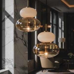 May include: Two pendant lights featuring a stacked design with a smaller, opaque white glass orb resting atop a larger, transparent amber-tinted glass bowl. Each fixture is suspended by a thin black cord and includes a metallic gold-toned connector.