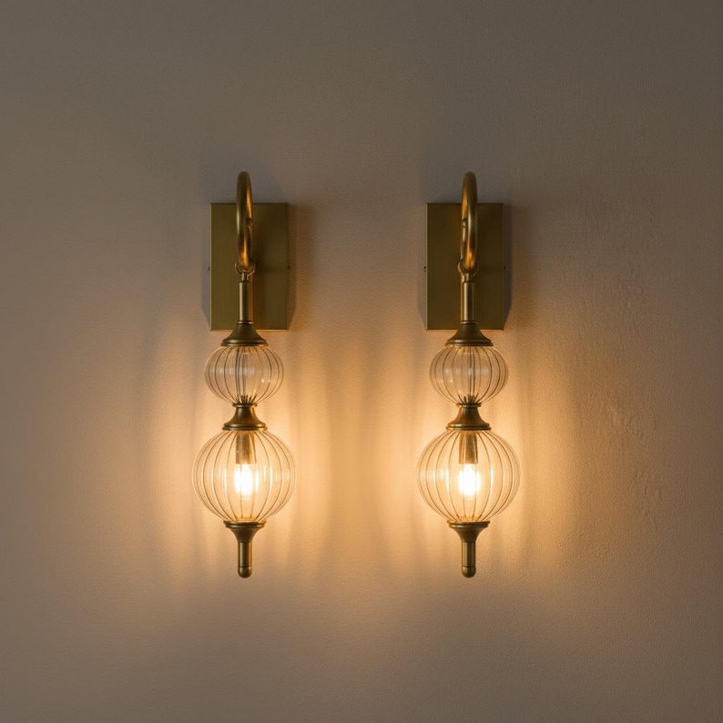 May include: Two wall sconces with a brass finish. Each sconce features a rectangular backplate, a curved arm, and two clear, ribbed glass globes. The sconces are illuminated, casting a warm glow.