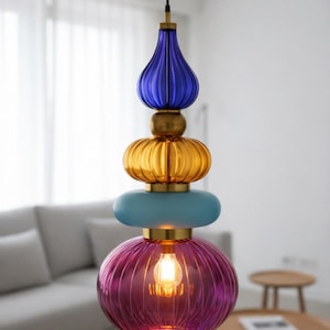 Puede incluir: Una lámpara colgante de cristal multicolor. La lámpara tiene un diseño en capas con elementos de vidrio azul, dorado y rosa. Cuelga de un cable negro y contiene una bombilla cálida. Un accesorio decorativo para el hogar.