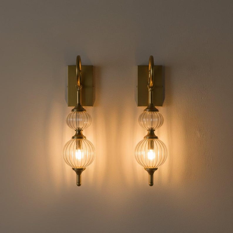 May include: Two wall sconces with a brass finish. Each sconce features a rectangular backplate, a curved arm, and two clear, ribbed glass globes. The sconces are illuminated, casting a warm glow.