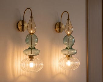 May include: Two decorative wall sconces with a brass finish. Each sconce features a clear, ribbed glass bottom, a teal-colored glass center, and a light amber glass top. The sconces are mounted on a light-colored wall.