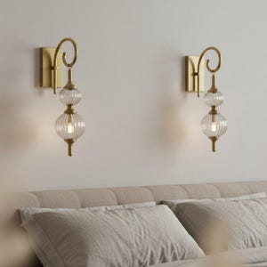 May include: Two gold-toned wall sconces with clear, ribbed glass globes and a decorative swirl design. The sconces are mounted on a light-colored wall above a bed with neutral-colored bedding. The sconces provide soft lighting.