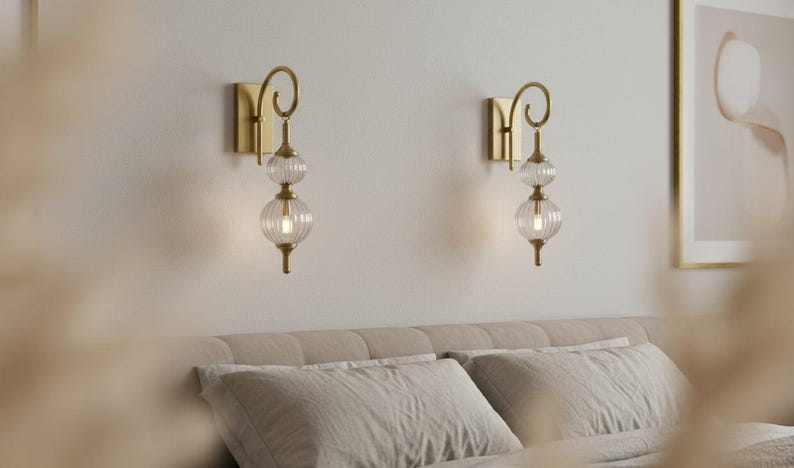 May include: Two gold-toned wall sconces with clear, ribbed glass globes and a decorative swirl design. The sconces are mounted on a light-colored wall above a bed with neutral-colored bedding. The sconces provide soft lighting.