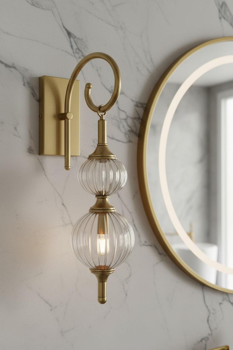 May include: A gold-toned wall sconce with a curved arm and two clear, ribbed glass globes. The fixture is mounted on a rectangular base and hangs near a round mirror with a gold frame. The light fixture is designed for indoor use.