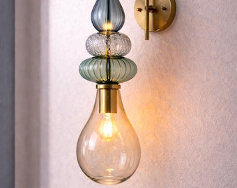 May include: A wall sconce featuring a brass finish arm and circular mounting plate. The fixture includes three stacked glass elements in teardrop and disc shapes with blue and clear textures, ending in a large clear glass bulb with a warm glow.
