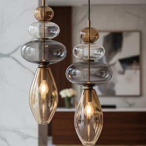 May include: Two modern pendant lights with a stacked glass design. Each light features a gold-toned top sphere, clear and gray glass elements, and a teardrop-shaped amber glass bottom. The lights hang from black cords.