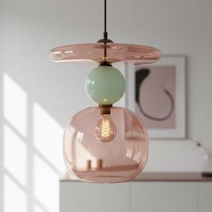 May include: A pendant light fixture featuring a flat, translucent pink glass disc at the top, a central mint green glass sphere, and a large, rounded, translucent pink glass shade at the bottom, all suspended by a thin black cord.