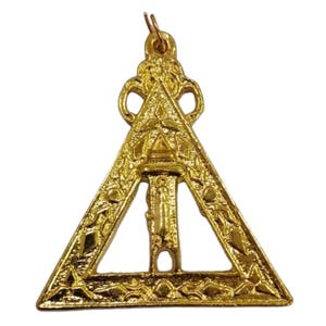 May include: A gold-colored pendant in the shape of a triangle with ornate detailing. The pendant features a central vertical design and decorative patterns along the edges. It has a loop at the top for hanging.
