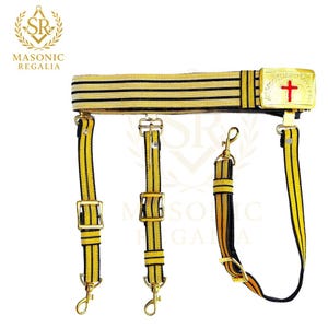 May include: Masonic Regalia belt with gold and black stripes, featuring a gold buckle with a red cross. The belt has three suspender straps with gold hardware and clasps. The Masonic Regalia logo is in the upper left corner.