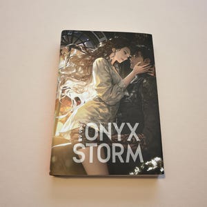 May include: A book cover featuring a romantic illustration of a man and woman embracing. The woman has long, flowing hair and is wearing a light-colored dress. The man is in a dark jacket. The title "ONYX STORM" is prominently displayed.