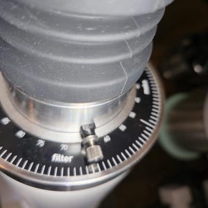 May include: Close-up of a coffee grinder with a black and silver design. The grinder features a dial with numbers and the word "filter". The top portion is a ribbed, dark grey material.