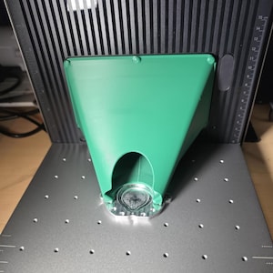 Op de afbeelding: Een groen, trechtervormig 3D-geprint onderdeel met een brede bovenkant en een smalle, boogvormige opening aan de basis. Het is gemonteerd op een zilveren, ronde metalen plaat met een gestructureerd, concentrisch cirkelvormig patroon.