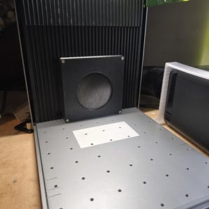 May include: A grey metal machine featuring a black, ridged back panel and a square, black component with a circular aperture. The machine has a flat, grey surface with evenly spaced holes and a white square sticker.