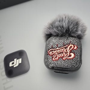 Pode incluir: Um transmissor de microfone DJI quadrado preto e um receptor de microfone quadrado preto com um protetor de vento cinzento felpudo. O receptor apresenta uma superfície preta texturizada com o texto vermelho brilhante Pixel 2 Plastics.