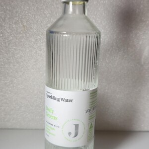 May include: A clear glass bottle of Daily Greens sparkling water with a black cap and two black bottle stoppers. The bottle has a white label with green text. The bottle stoppers are cylindrical with a handle.