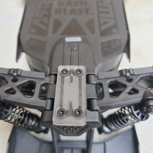 Peut inclure: Châssis de voiture télécommandée noir et gris avec ressorts de suspension et le texte "DON'T JUST BASH. BLAST.". Le châssis comporte un composant en plastique transparent avec quatre vis.