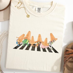 Puede incluir: Camiseta de color marfil con un gr&aacute;fico de personajes de dibujos animados cruzando un paso de cebra a rayas blancas y negras, con conos de tr&aacute;fico naranjas. La camiseta se combina con sandalias marrones y un sombrero beige.