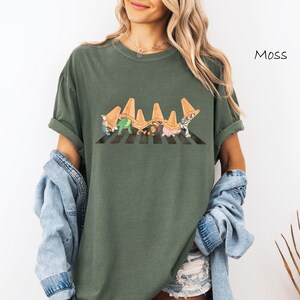 Puede incluir: Camiseta verde musgo con un gr&aacute;fico caprichoso de personajes de dibujos animados cruzando un paso de cebra a rayas blancas y negras. La palabra "Moss" est&aacute; impresa en la parte superior derecha. La camiseta tiene mangas cortas y un corte holgado.