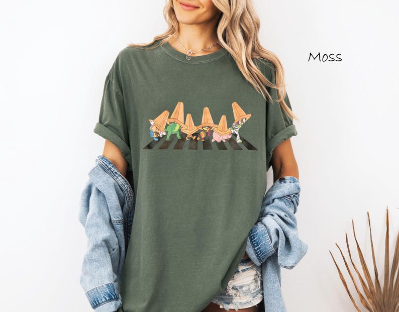 Puede incluir: Camiseta verde musgo con un gr&aacute;fico caprichoso de personajes de dibujos animados cruzando un paso de cebra a rayas blancas y negras. La palabra "Moss" est&aacute; impresa en la parte superior derecha. La camiseta tiene mangas cortas y un corte holgado.