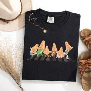 Puede incluir: Camiseta negra con un dise&ntilde;o de dibujos animados de personajes cruzando un paso de cebra, cada uno llevando un cono de tr&aacute;fico naranja. La camiseta se combina con sandalias marrones, un collar dorado y un sombrero color crema con una banda marr&oacute;n.