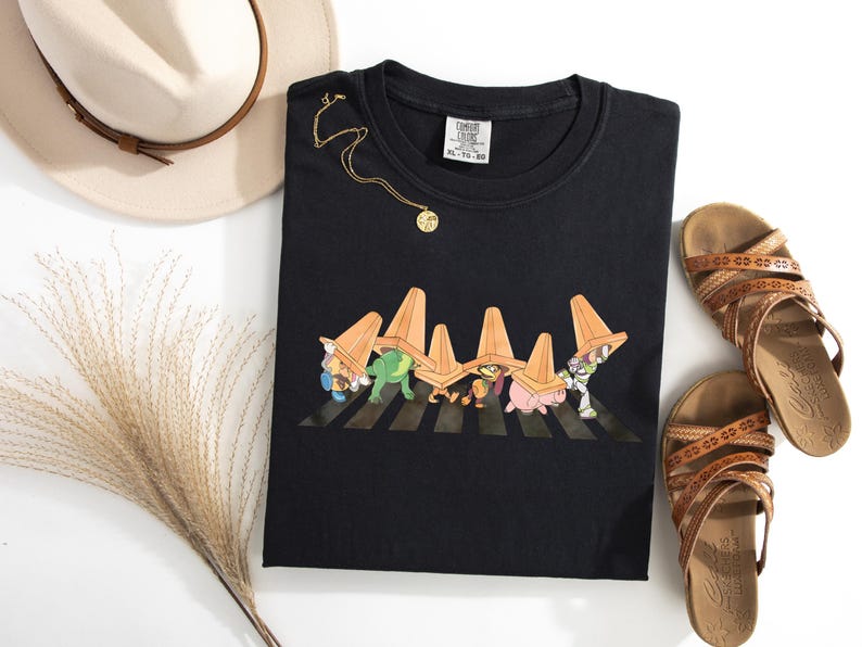 Puede incluir: Camiseta negra con un dise&ntilde;o de dibujos animados de personajes cruzando un paso de cebra, cada uno llevando un cono de tr&aacute;fico naranja. La camiseta se combina con sandalias marrones, un collar dorado y un sombrero color crema con una banda marr&oacute;n.