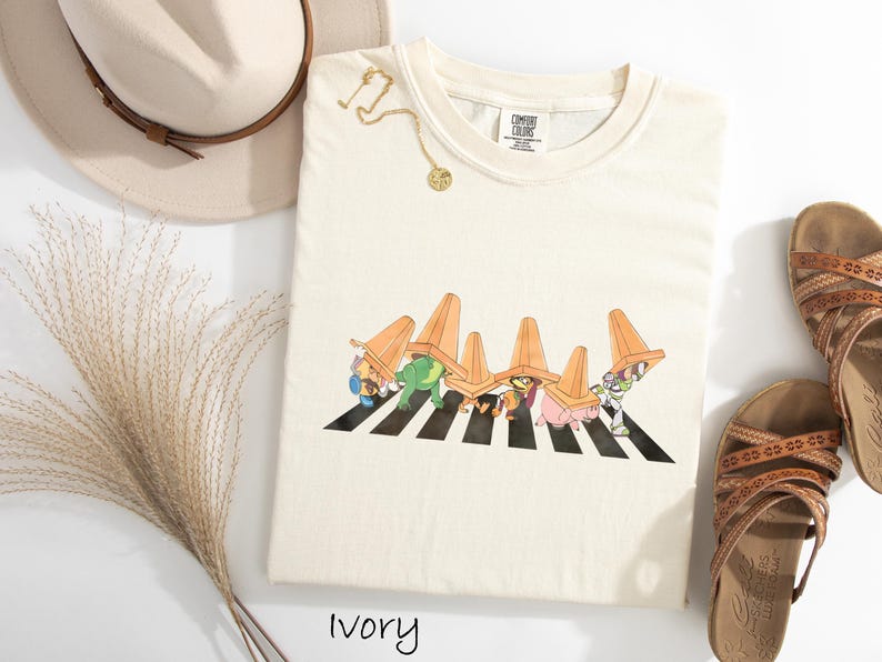 Puede incluir: Camiseta de color marfil con un gr&aacute;fico de personajes de dibujos animados cruzando un paso de cebra a rayas blancas y negras, con conos de tr&aacute;fico naranjas. La camiseta se combina con sandalias marrones y un sombrero beige.