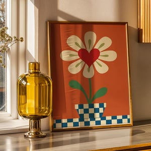 Puede incluir: Una impresión artística enmarcada con una flor con un corazón rojo en el centro y pétalos blancos sobre un fondo naranja. Un jarrón marrón con flores secas, una botella de vidrio amarilla, un cilindro blanco y un recipiente negro están sobre una superficie de madera.
