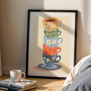 Op de afbeelding: Een ingelijste kunstdruk met een stapel van vijf kleurrijke theekopjes met bloemmotieven. De theekopjes zijn in tinten geel, groen, blauw, rood en blauw, met een bijpassende schotel aan de onderkant. Een witte mok en boeken staan op de voorgrond.