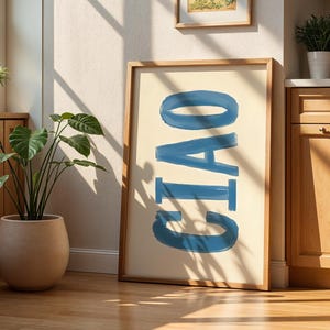 Op de afbeelding: Een ingelijste kunstprint met het woord "CIAO" in blauwe penseelstreken op een crèmekleurige achtergrond. Het kunstwerk leunt tegen een muur in een kamer met houten kasten, een plant in een pot en een raam. De kamer wordt verlicht door natuurlijk licht.