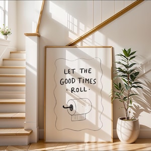 Puede incluir: Un cuadro enmarcado con el texto "LET THE GOOD TIMES ROLL" y un dibujo de un rollo de papel higiénico. La obra de arte está en un marco de color claro y se exhibe en un interior moderno y luminoso con una escalera y una planta en maceta.